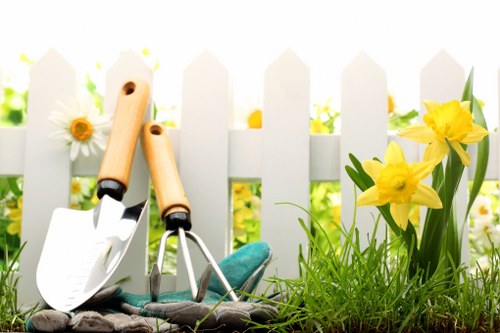 The image depicts a small section of a garden with a white picket fence in the background, characteristic of a suburban outdoor space in the UK. In the foreground, there is a patch of vibrant green grass, well-maintained and lush, with some soil visible alongside it. To the right, a group of blooming yellow flowers, likely daffodils, are growing from the soil bed, adding natural colour and variety to the garden area. On the left, two gardening toolsâ€”a trowel and a hand rakeâ€”rest against the fence, suggesting recent or ongoing gardening activity. The tools have wooden handles and metal working ends, and they are positioned on a textured, dark fabric or mat that is likely used to keep the tools clean or organized. The overall scene is illuminated by natural daylight, indicating a bright, clear day suitable for outdoor gardening work. This setting reflects typical garden maintenance activities and highlights the importance of outdoor spaces in residential properties in Brimsdown, aligning with professional gardening services offered in the area, such as lawn care, flower bed maintenance, and planting.